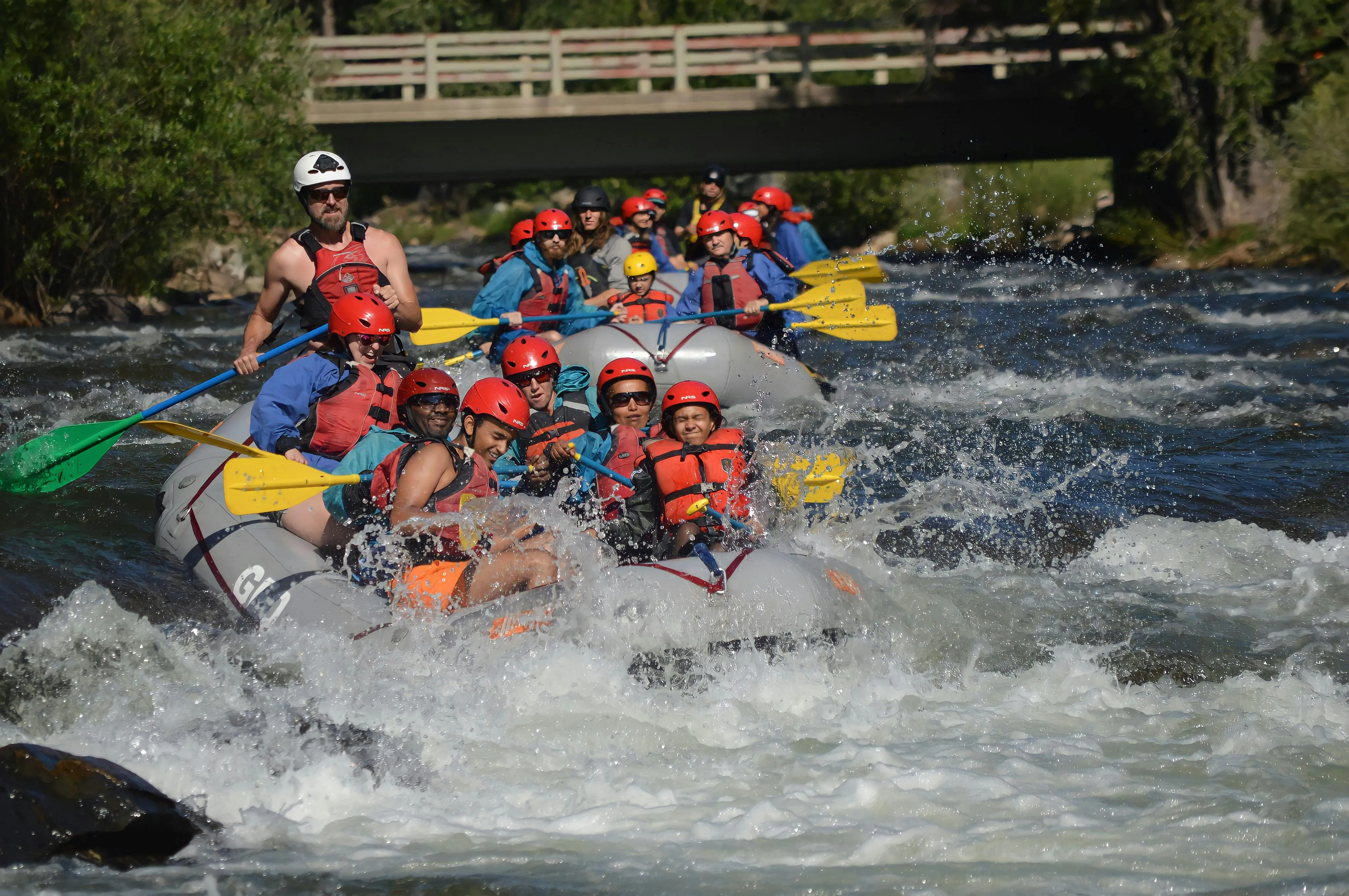 Group of people in an inflatable raft navigating turbulent whitewater rapids wearing helmets and life jackets led by a standing guide; splashing river water with rocky banks and a wooden bridge in the background; energetic and exhilarated mood